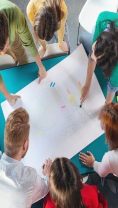 Um grupo multiétnico de pessoas está sentado ao redor de uma mesa de trabalho. No centro, está posicionada uma cartolina branca. Várias pessoas escrevem sobre ela, usando como facilitador a Metodologia Ágil.