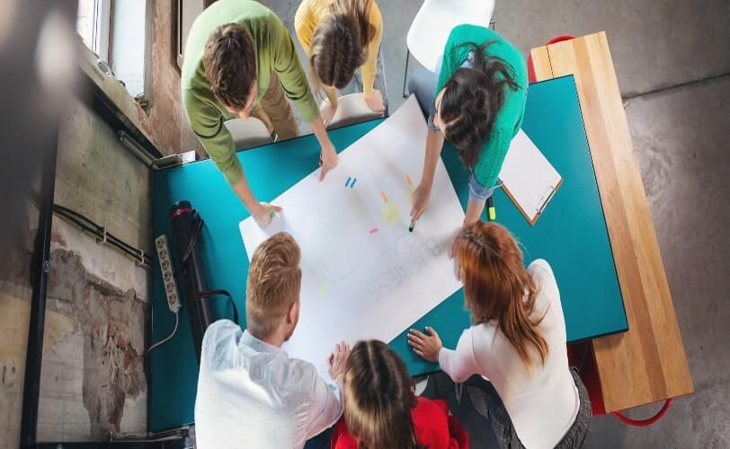 Um grupo multiétnico de pessoas está sentado ao redor de uma mesa de trabalho. No centro, está posicionada uma cartolina branca. Várias pessoas escrevem sobre ela, usando como facilitador a Metodologia Ágil.