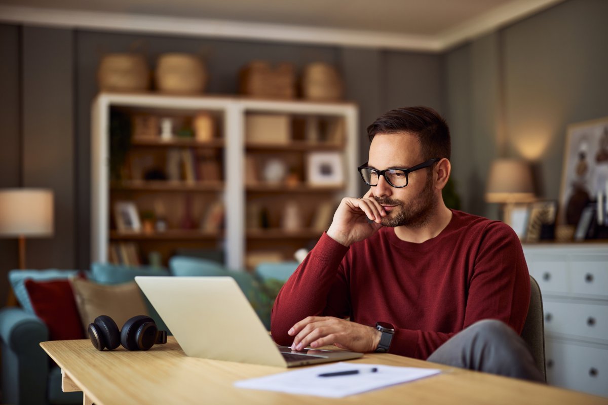 A imagem mostra um homem trabalhando no que parece ser a sala de estar de uma casa. Ele está olhando para o notebook com uma expressão pensativa, analisando os principais pontos entre GraphQL vs REST.