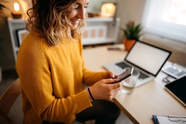 A imagem mostra a mulher em um escritório, aparentemente em sua casa. Há um notebook ligado sobre a mesa. Ela mexe no celular para verificar um e-mail marketing enviado por uma empresa.