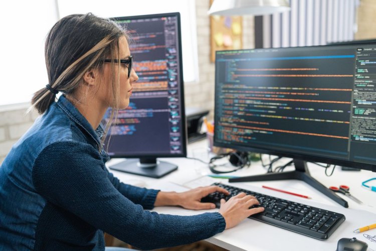 A imagem mostra uma mulher branca de óculos em um escritório. Ela está sentada diante de dois monitores, lendo várias linhas de código de programação, que possivelmente serão usadas com uma API para desenvolvedores. 