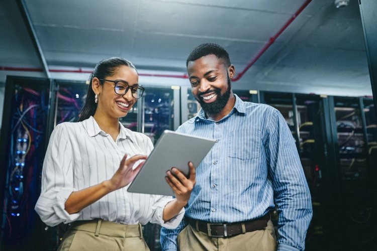 A imagem mostra um homem e uma mulher conversando no que parece ser a sala de um servidor de uma empresa. Ela segura um tablet e aponta para a tela, possivelmente comentando com o colega sobre as vantagens da migração para nuvem.