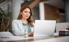 A imagem mostra uma mulher com roupas sociais usando um notebook no que parece ser um escritório. Ela aparenta ter um olhar confiante e seguro, pois está acessando um site que utiliza Wildcard SSL para aumentar a segurança dos dados.