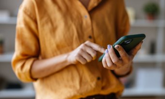 A imagem mostra uma mulher com um celular na mão. O rosto dela não aparece. Ela toca na tela com o dedo indicador, possivelmente em um botão de CTA para realizar uma compra ou outra ação com foco em conversão.