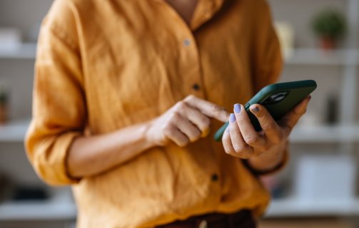 A imagem mostra uma mulher com um celular na mão. O rosto dela não aparece. Ela toca na tela com o dedo indicador, possivelmente em um botão de CTA para realizar uma compra ou outra ação com foco em conversão.