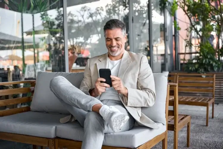 A imagem mostra um homem sorrindo enquanto mexe em seu smartphone. Ele está em uma área de descanso e, possivelmente, foi impactado de maneira positiva pela mensagem de alguma empresa que usou um call to action correto. 