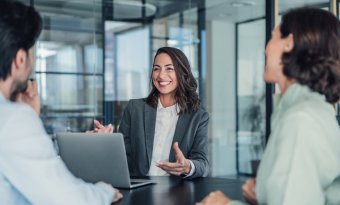 A imagem mostra três pessoas conversando no que parece ser uma reunião em uma empresa. A mulher em destaque está na frente de um notebook e gesticula para explicar o processo de como criar uma marca do zero.