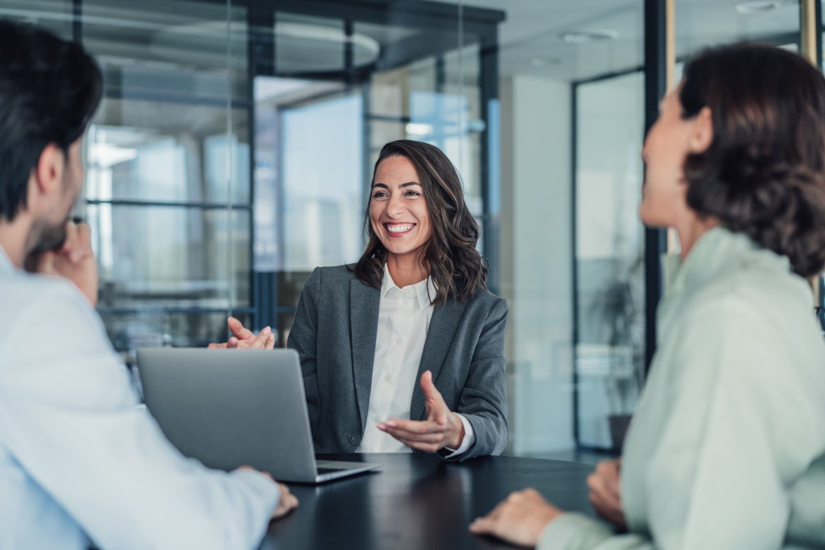 A imagem mostra três pessoas conversando no que parece ser uma reunião em uma empresa. A mulher em destaque está na frente de um notebook e gesticula para explicar o processo de como criar uma marca do zero.