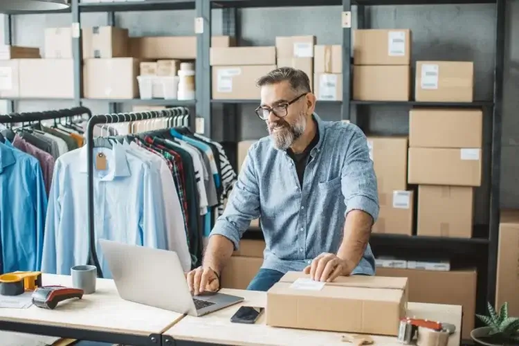 A imagem mostra um homem, que certamente é dono de uma loja de roupas que vende pela internet, pois há algumas araras com camisas penduradas e caixas de papelão ao seu redor. Ele está digitando em um notebook para finalizar o processo de criação da sua marca. 