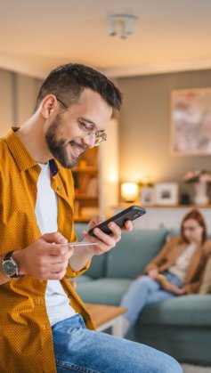 A imagem mostra um homem branco segurando um celular. Ele está fazendo uma compra na sala da casa dele, segurando um cartão de crédito para digitar os dados necessários e concluir a transação.