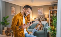 A imagem mostra um homem branco segurando um celular. Ele está fazendo uma compra na sala da casa dele, segurando um cartão de crédito para digitar os dados necessários e concluir a transação.