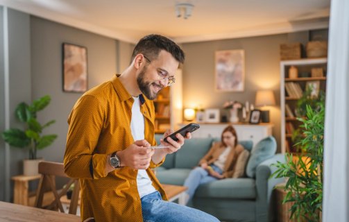 A imagem mostra um homem branco segurando um celular. Ele está fazendo uma compra na sala da casa dele, segurando um cartão de crédito para digitar os dados necessários e concluir a transação.