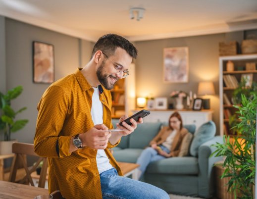 A imagem mostra um homem branco segurando um celular. Ele está fazendo uma compra na sala da casa dele, segurando um cartão de crédito para digitar os dados necessários e concluir a transação.