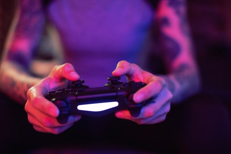 A imagem mostra uma pessoa com braços tatuados jogando videogame. O rosto dela não aparece. Ela está segurando um controle de PlayStation 5, representando a paixão por jogos entre aqueles que são amantes de novas tecnologias. 