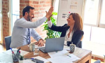 a imagem mostra um executivo e uma executiva felizes em um ambiente semelhante a um escritório. Os dois estão fazendo um high five. A imagem faz referência ao sucesso em comunicação empresarial abordado no conteúdo.