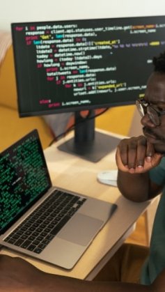 Um jovem desenvolvedor afro-americano está concentrado, analisando um código-fonte em linguagem Go exibido em duas telas de computador. Ele está sentado em uma mesa de trabalho. O ambiente é organizado, com uma decoração aconchegante ao fundo.