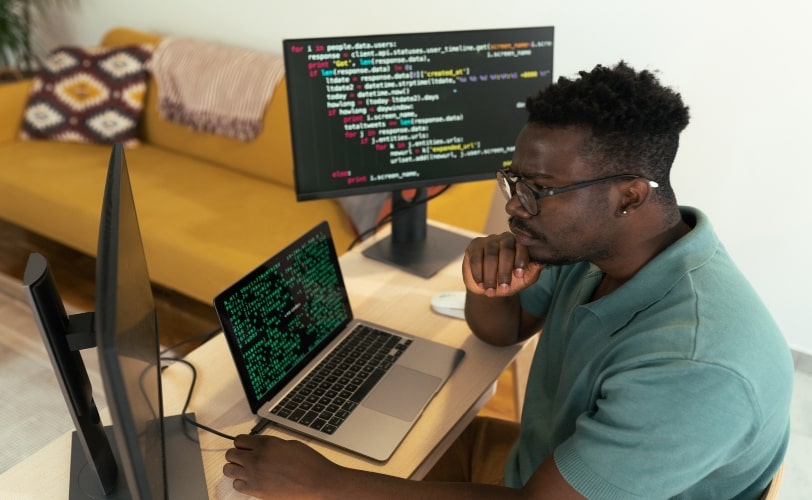 Um jovem desenvolvedor afro-americano está concentrado, analisando um código-fonte em linguagem Go exibido em duas telas de computador. Ele está sentado em uma mesa de trabalho. O ambiente é organizado, com uma decoração aconchegante ao fundo.