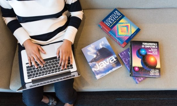 Na imagem temos a visão aérea de uma pessoa sentada em um sofá com uma notebook no colo e ao seu lado há três livros sobre desenvolvimento web.