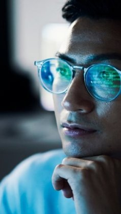 A imagem mostra um homem olhando para uma tela de computador. Ele está programando com o cURL, por isso é possível ver alguns códigos refletidos na cor verde nas lentes de seus óculos.