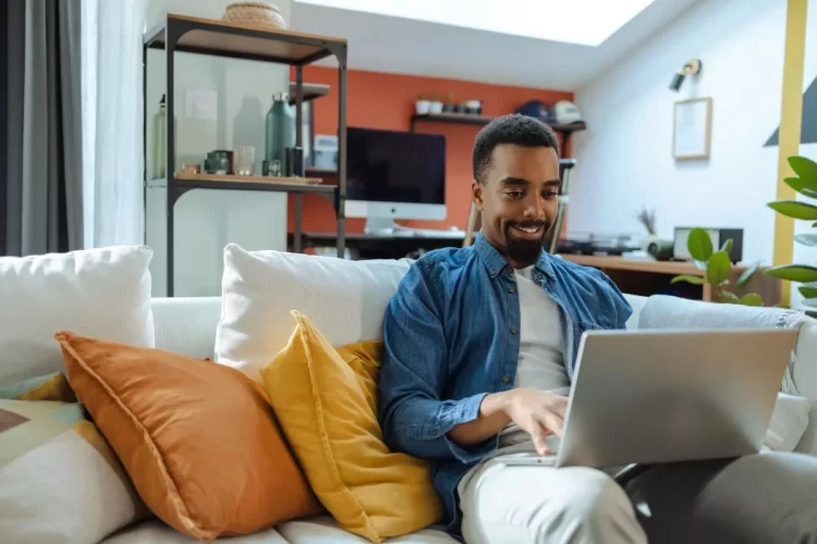 A imagem mostra um homem negro utilizando a internet para saber tudo sobre Inteligência Artificial. Ele está em casa para realizar essa pesquisa, sentado em seu sofá, usando um notebook.