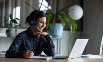 A imagem mostra uma mulher sentada a uma mesa de trabalha, em frente a seu notebook e com fones de ouvidos. Ela está sorrindo.