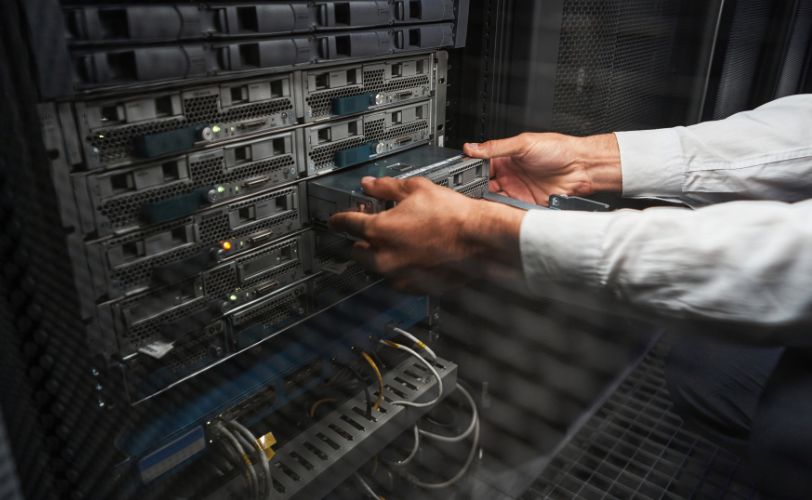 Técnico instalando ou ajustando um servidor em um data center empresarial. O rosto dele não aparece, apenas as mãos segurando o que parece ser um módulo.