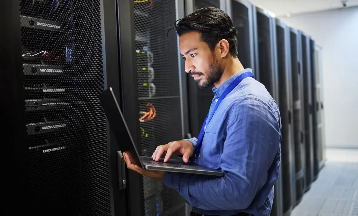 A imagem mostra um homem dentro da sala de servidores de uma empresa. Ele está digitando no teclado de um notebook, possivelmente realizando algumas configurações de servidor proxy.