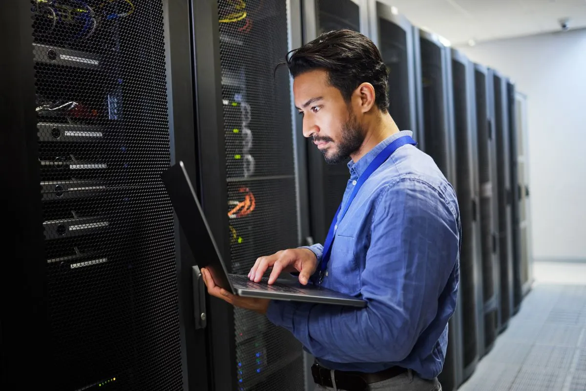 A imagem mostra um homem dentro da sala de servidores de uma empresa. Ele está digitando no teclado de um notebook, possivelmente realizando algumas configurações de servidor proxy.