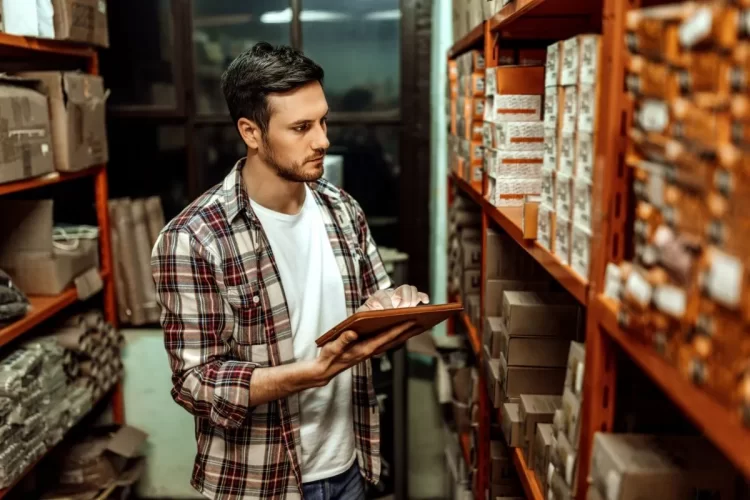 A imagem mostra um homem segurando um tablet. Ele está analisando os produtos que estão em um estoque e, possivelmente, verificando os SKUs de cada um deles para fazer anotações.