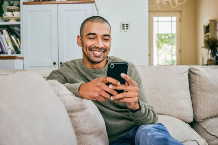 A imagem mostra um homem sentado no sofá de sua casa, olhando para um smartphone e sorrindo. Ele está digitando algo na tela, o que indica que possivelmente está interagindo com alguma publicação no Instagram.