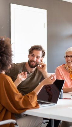 A imagem apresenta um trio multiétnico de pessoas, sendo uma delas afro-americana, que está de costas para a imagem, conversando com um homem branco e jovem e uma mulher branca de meia idade.