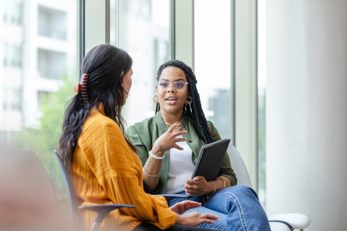 A imagem apresenta duas mulheres sentadas em um sofá em uma empresa, sendo que uma delas está de costas. A outra, de frente, é uma mulher jovem e afro-americana, segurando um notebook em uma das mãos. Ambas vestem roupas casuais e conversam entre si. 