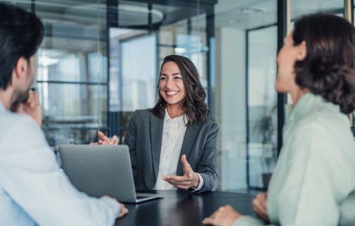 A imagem mostra uma mulher conversando com duas pessoas. Ela está em um escritório, possivelmente aplicando conceitos de marketing pessoal para destacar as habilidades dela.