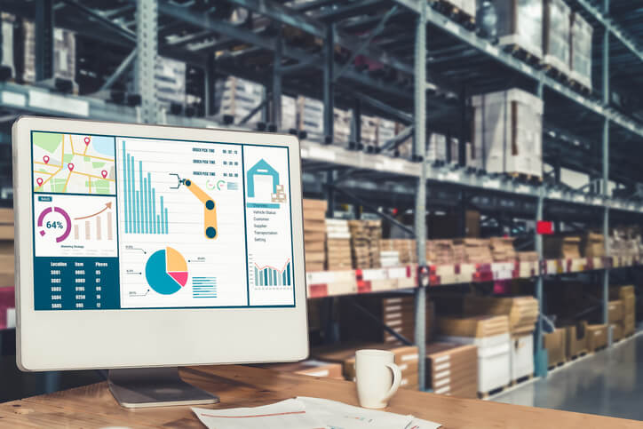 Uma tela de computador está em primeiro plano exibindo um dashboard com vários gráficos, tabelas e análises de dados. No fundo, há prateleiras cheias de caixas e pacotes em um depósito. Uma xícara e papéis também estão na mesa.