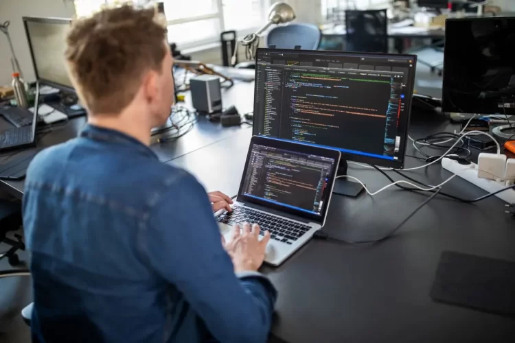 A imagem mostra um homem em um escritório. Ele está diante de um notebook, olhando para uma tela com várias linhas de código, todas geradas após aplicar algumas ideias de lógica de programação. 