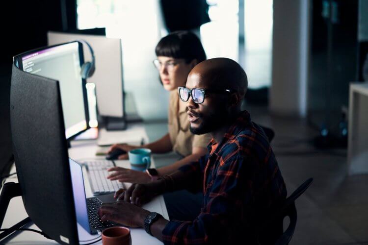 A imagem mostra uma dupla de desenvolvedores, um homem negro e uma mulher branca, trabalhando dentro de uma empresa. Cada um está sentado na frente de um monitor, possivelmente analisando a estrutura de códigos gerada com o auxílio do Domain-Driven Design. 