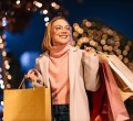 A imagem mostra uma mulher segurando algumas sacolas de compra. Ela provavelmente adquiriu presentes para o Natal, pois ao fundo há algumas árvores com diversas luzinhas piscando.