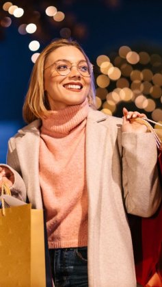 A imagem mostra uma mulher segurando algumas sacolas de compra. Ela provavelmente adquiriu presentes para o Natal, pois ao fundo há algumas árvores com diversas luzinhas piscando.