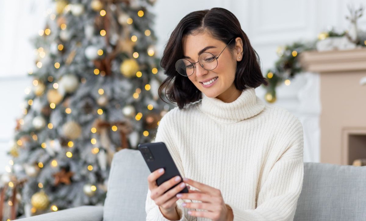 A imagem mostra uma mulher de óculos sentada na sala de uma casa. Há uma árvore de Natal enfeitada e iluminada ao fundo. Ela segura o celular e sorri enquanto lê uma mensagem de Natal enviada por uma empresa.