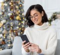 A imagem mostra uma mulher de óculos sentada na sala de uma casa. Há uma árvore de Natal enfeitada e iluminada ao fundo. Ela segura o celular e sorri enquanto lê uma mensagem de Natal enviada por uma empresa.