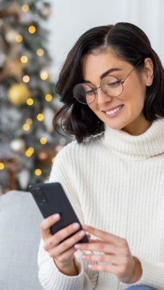 A imagem mostra uma mulher de óculos sentada na sala de uma casa. Há uma árvore de Natal enfeitada e iluminada ao fundo. Ela segura o celular e sorri enquanto lê uma mensagem de Natal enviada por uma empresa.