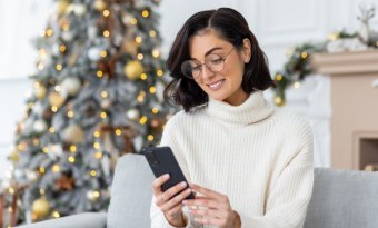 A imagem mostra uma mulher de óculos sentada na sala de uma casa. Há uma árvore de Natal enfeitada e iluminada ao fundo. Ela segura o celular e sorri enquanto lê uma mensagem de Natal enviada por uma empresa.