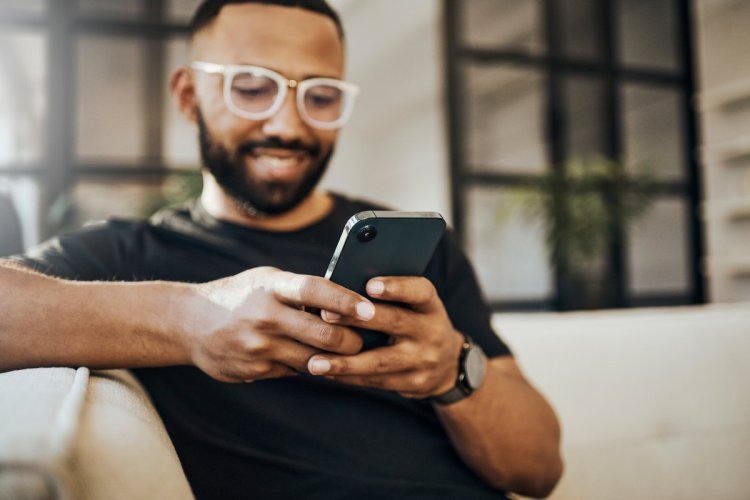 A imagem mostra um homem sentado no sofá do que parece ser a casa dele. Ele está lendo algo em seu smartphone e sorrindo, possivelmente feliz por ter recebido uma mensagem de Natal.