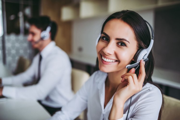 Mulher sorridente usando fone de ouvido, trabalhando em um ambiente de atendimento ao cliente ou call center, com um colega ao fundo.
Legenda: Empatia no atendimento transforma relações! Coloque-se no lugar do cliente e faça a diferença com um serviço humano e ágil.