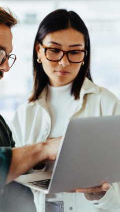 A imagem mostra um homem e uma mulher conversando em um escritório. Ele está segurando um notebook e apontando algo para a tela, possivelmente dando a ela dicas de como saber se um site é seguro.