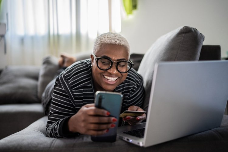 A imagem mostra uma mulher fazendo compras em uma loja online. Ela está em sua casa, aparentemente na sala, e vai finalizar pagando com o cartão de crédito em seu celular, após verificar que o site que está acessando é seguro. 