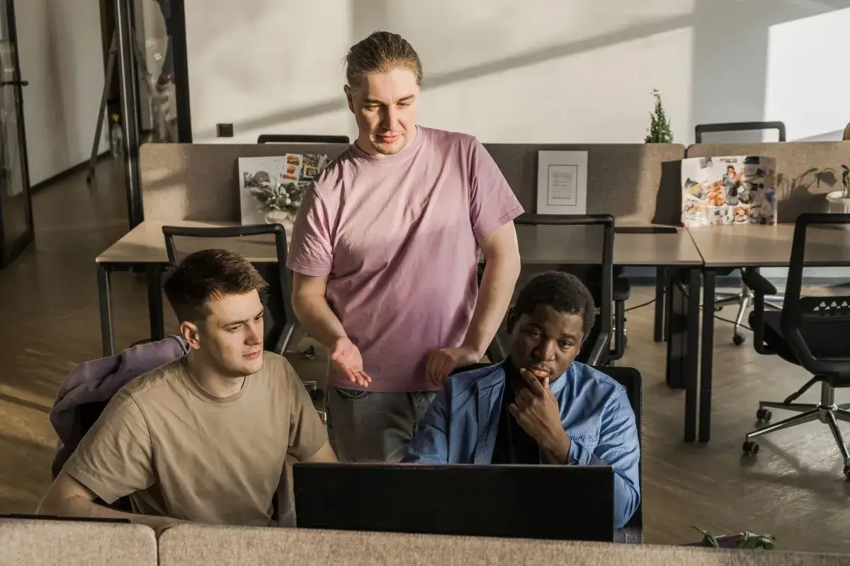 Três homens, dois brancos e um negro, conversando em um espaço de coworking, enquanto programam usando laptop. Um está olhando para a própria tela, enquanto os outros dois olham para a tela do primeiro. Um está de pé, os outros dois estão sentados em cadeiras de escritório.