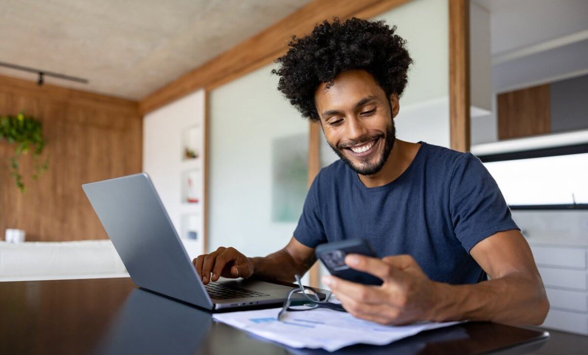 Homem negro sentado diante de um laptop. Ele observa o celular e sorri. Há um par de óculos e papéis em cima da mesa. O ambiente é clean, com detalhes em madeira.