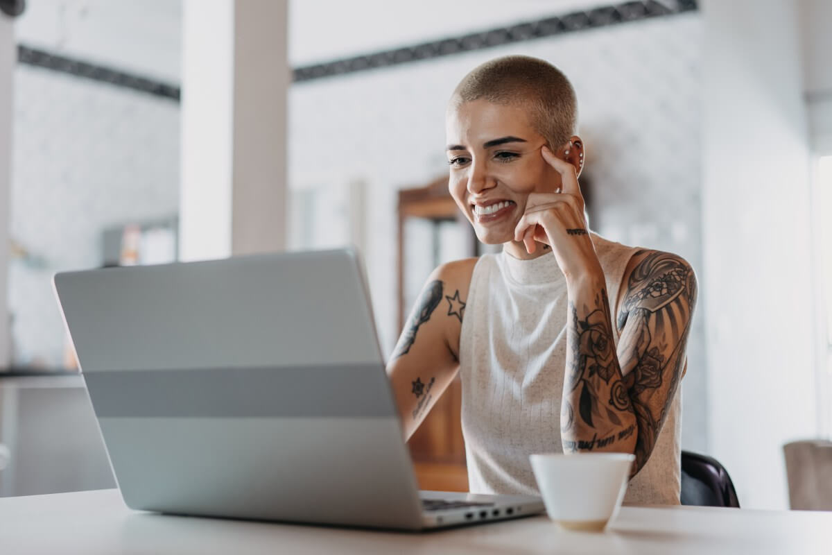 Mulher careca sorri diante de computador. Há uma xícara de café ao lado do computador. O ambiente é claro. 