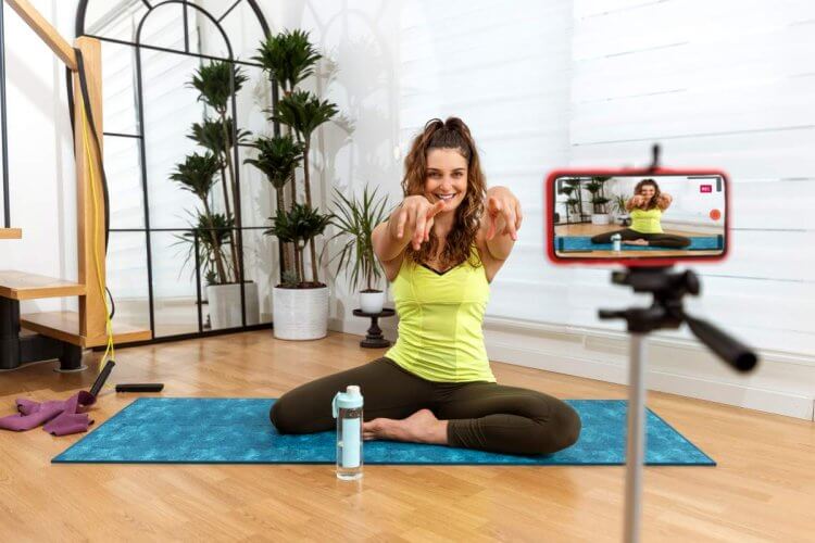 Mulher parece dar aulas de yoga via internet. Ela está sentada no chão, sobre um tapete e com uma garrafa de água por perto. Um celular apoiado em um tripé filma os seus movimentos. 